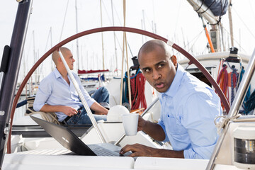 man working at the laptop and drinking coffee and watching passers by on private yachts in the...
