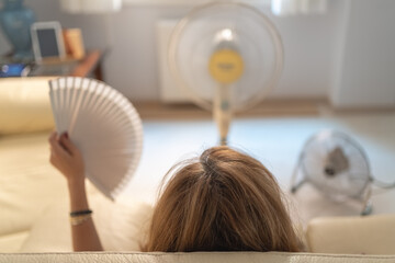 Woman sitting on a sofa with several fans giving her air and a fan in her hand, high summer...