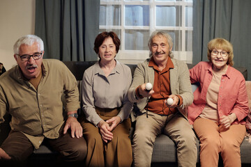 Group of cheerful mature friends sitting on sofa in living room in front of tv set while playing video game and commenting their actions