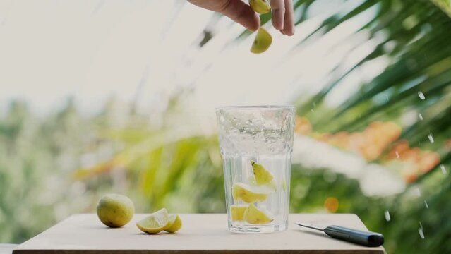 Close a glass of water , lime and knife on a wooden table.  A man's hand throwing lime slices into a glass of water . In the background, palm leaves and the sea on a sunny summer day. Slow motion.
