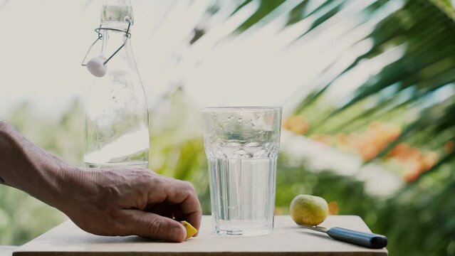 Close a glass of water , lime and knife on a wooden table.  A man's hand throwing lime slices into a glass of water . In the background, palm leaves and the sea on a sunny summer day. Slow motion.