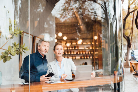 Businessman holding mobile phone standing by businesswoman in cafe seen through window