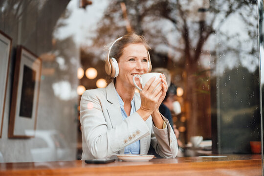 Smiling businesswoman holding coffee cup listening music through wireless headphones in cafe