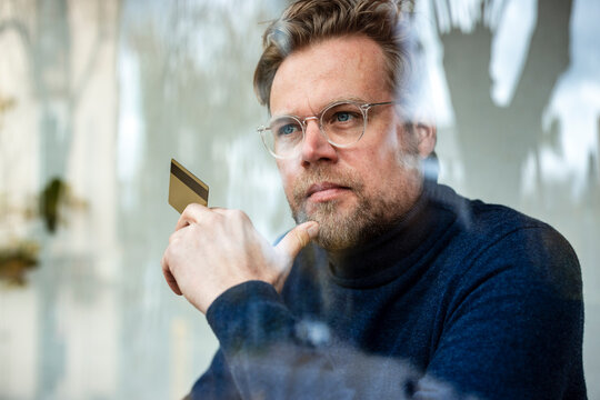 Thoughtful businessman with holding credit card in cafe seen through glass window
