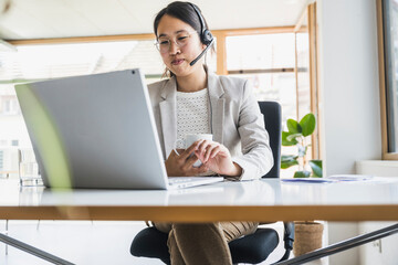 Smiling businesswoman with headset working on laptop at desk