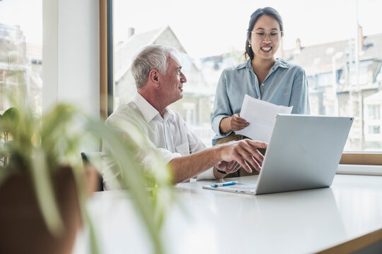 Happy businessman discussing work with colleague standing by desk in office