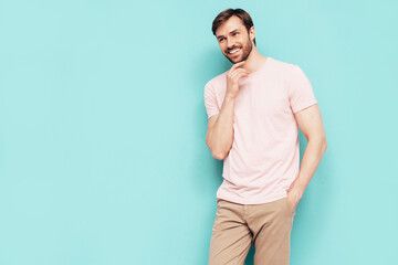 Fototapeta na wymiar Portrait of handsome smiling stylish hipster lambersexual model. Sexy man dressed in pink T-shirt and trousers. Fashion male isolated on blue wall in studio