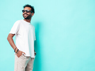 Handsome smiling hipster model. Sexy unshaven man dressed in white summer t-shirt and jeans clothes. Fashion male posing near blue wall in studio. Isolated. In sunglasses