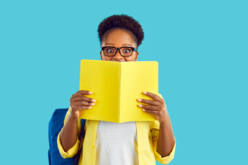 Fototapeta Happy cheerful smart Afro American high school, college or university student girl in eyeglasses isolated on blue background hides half of her face behind book or diary with notes and looks at camera obraz