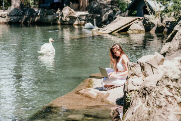 a beautiful woman in a long white dress sits near a lake with swans and works at a laptop