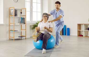 Professional male physiotherapist works with senior female patient in rehabilitation center. Smiling elderly woman sitting on fitness ball in medical office and doing exercises with resistant band.