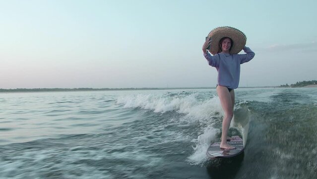 A surfer in a straw hat jumps on a wakeboard and takes a selfie on her phone. An experienced wakeboarder sprays water drops into the camera.