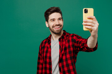 Young happy smiling fun caucasian man he 20s wearing red shirt grey t-shirt doing selfie shot on mobile cell phone post photo on social network isolated on plain dark green background studio portrait