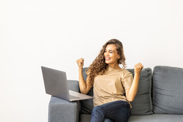Emotional young woman with laptop celebrating victory on sofa at home