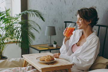 Side view young smiling woman wear white shirt pajama she lying in bed eat breakfast drink orange...