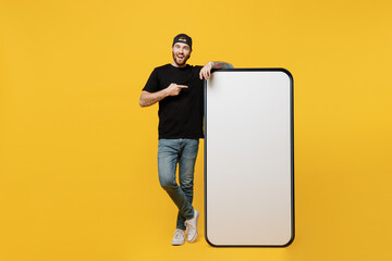 Full body young tattooed man he wears black t-shirt cap point index finger on big huge blank screen mobile cell phone workspace mockup area isolated on plain yellow background Tattoo translate fun - obrazy, fototapety, plakaty