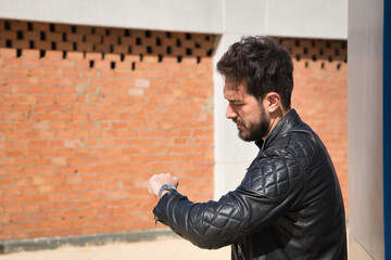 Handsome young man with beard, leather jacket, looking at the time on his luxury wristwatch. Concept beauty, fashion, trend, modern, luxury, jewelry, time, watches.
