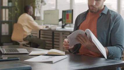 Slowmo of bearded young Caucasian marketing specialist reading and analyzing financial documents working in modern agency with African American colleague