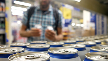 Close-up of many aluminium cans of beer or another drink on a store shelf and a male buyer takes a couple - obrazy, fototapety, plakaty