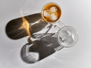 A glass thermo mug with ice tea and a mug with ice and sharp shadows stand on a white table