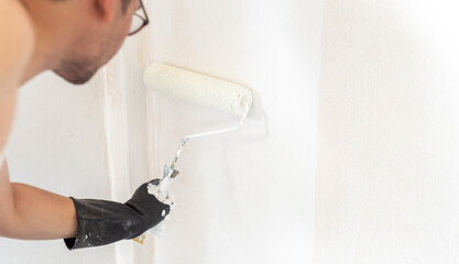 A white man is using a paint roller to paint to repair the walls of a room.