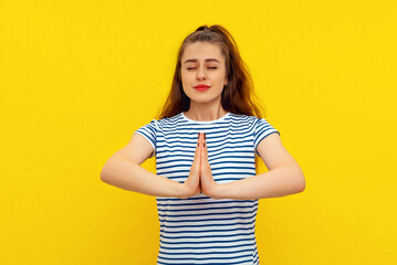 Calm beautiful young woman keeps palms pressed together, showing pray, namaste gesture, express gratitude, thank you sign, standing in white-blue striped t shirt over yellow background