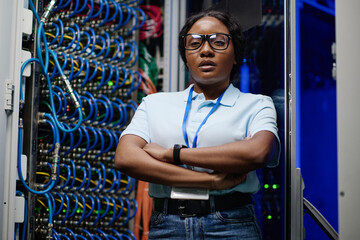 Portrait of African female IT engineer in eyeglasses standing with her arms crossed in data center and looking at camera