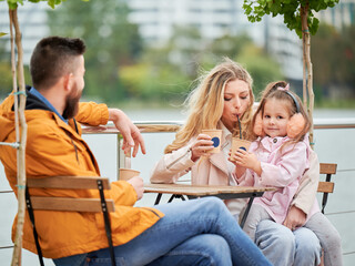 Young family man and woman drinking of coffee while spending time with daughter in outdoor cafe. Happy husband and wife sitting at the table with drink cups in new urban district.