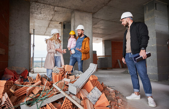 Man And Woman With Child Standing Inside Apartment Building Under Construction And Talking With Builder. Family Future Homeowners Discussing Building Process With Construction Worker.