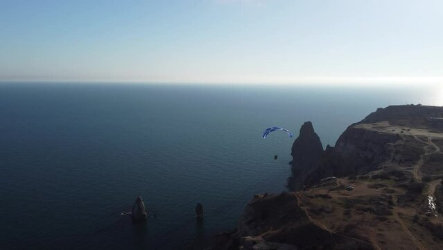 Aerial drone view of a man flying a white and blue paraglider over a hill and trees to the sea waves near the rocks. Active paraglider flight over the seascape with clear skies at suset. Extreme sport