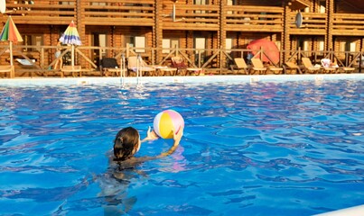 A girl in a bright bathing suit swims with an inflatable ball in a pool with clear water on a...