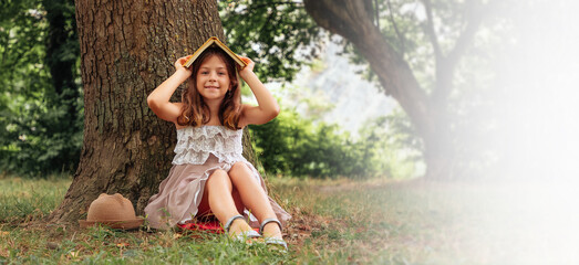 Cute schoolgirl girl in a straw hat and dress is sitting on the grass by a tree and made a house out of a book on her head. Copy space. School holidays. The concept of children's education