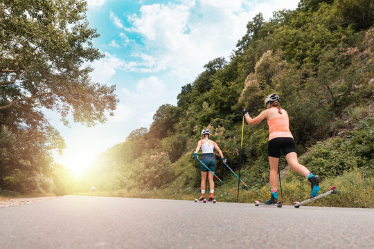 Two athletic women together training on the roller ski. Back low angle view. Copy space. Sunlight at finish. Concept of competition, biathlon, and summer workout