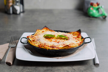 Traditional Lasagna made with minced beef bolognese topped with basil leafs, in the black rustic metal pot