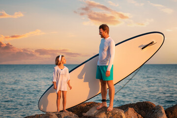 Summer activity vacations. Father and daughter standing with sup board at big beach rocks. Sunset...