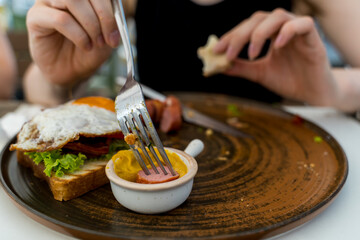 A close up girl is eating healthy English breakfast during the day in a restaurant cutting sausage and sandwich 