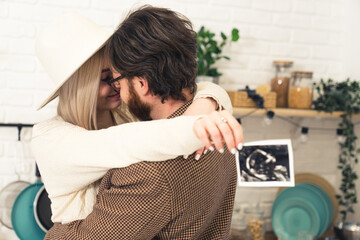 Caucasian blonde woman in white hat hugging her partner with dark hair and beard holding ultrasound...