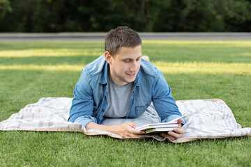 Young man relaxing and reading book while lying on lawn. Happy Man Lying On Green Grass and study remotely. Adult student reading in the park. Enjoying student life. smiling man working on outdoor