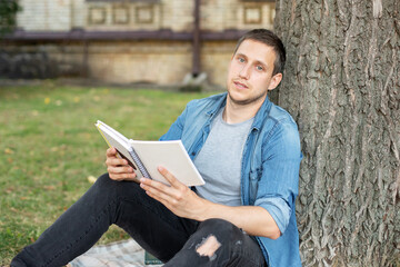 Young man relaxing and reading book while sit on lawn. Happy Man sitting On Green Grass and study remotely. Adult student reading in the park. Enjoying student life. smiling man working on outdoor 
