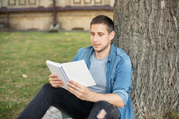 Obraz premium Young man relaxing and reading book while sit on lawn. Happy Man sitting On Green Grass and study remotely. Adult student reading in the park. Enjoying student life. smiling man working on outdoor 