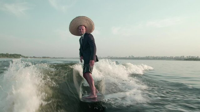A surfer jumping on a wakeboard in a suit with a straw hat on his head. An experienced wakeboarder sprays water drops into the camera.