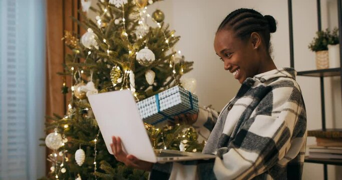 An African-looking girl received gift from boyfriend and has video chat. She is holding laptop and gift in hands. The girl is smiling and happy. She shook the box and guessed that in the middle.