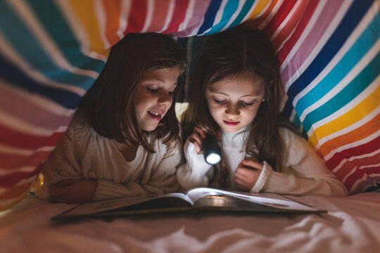 Two happy girls reading a book on the bed under the duvet using a torch light