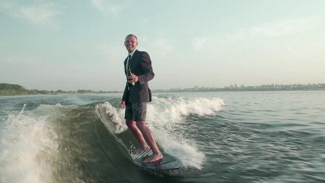A happy surfer jumping on a wakeboard in a suit with a bottle of champagne. An experienced wakeboarder sprays water drops into the camera.