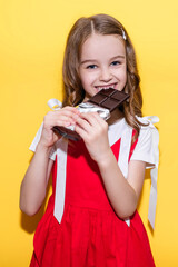 Naklejka premium Little girl bites a bar of chocolate on a yellow background. Happy child with face and hands covered in chocolate. The concept of children's happiness and love for sweets