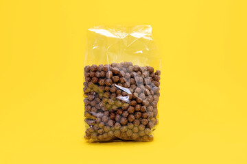 Round chocolate flakes in a transparent package on a yellow background. The packaged cereal food product is in the center of the image. Transparent packaging with round flakes in the form of balls