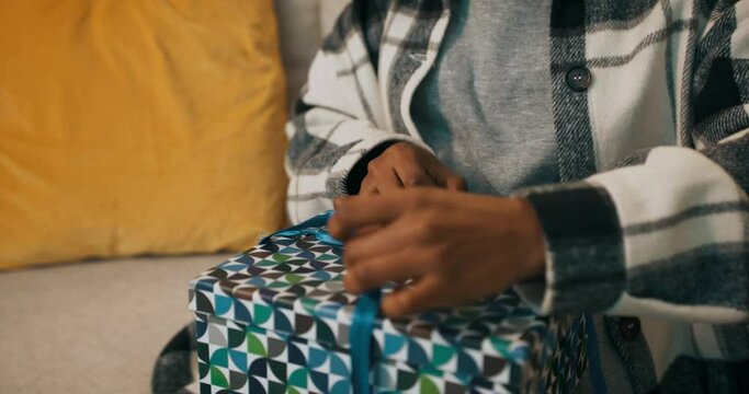 An African-looking girl dressed in checkered shirt sits on sofa and unpacks a gift. She unties the blue ribbon, opens the box and is upset by what she saw. She tossed the box aside in displeasure.