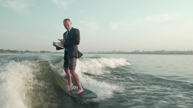 A surfer jumping on a wakeboard in a suit with a book in his hands takes photos of the pages of the book with his phone. An experienced wakeboarder sprays water drops into the camera.