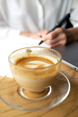 Beautiful business woman making notes on the background and transparent cup of coffee and laptop on the foreground