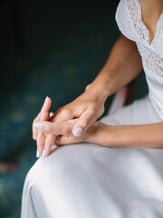 The bride in a silk dress with lace sits holding her hand in her hand. On the hands of a wedding nude manicure, wedding, and engagement rings.